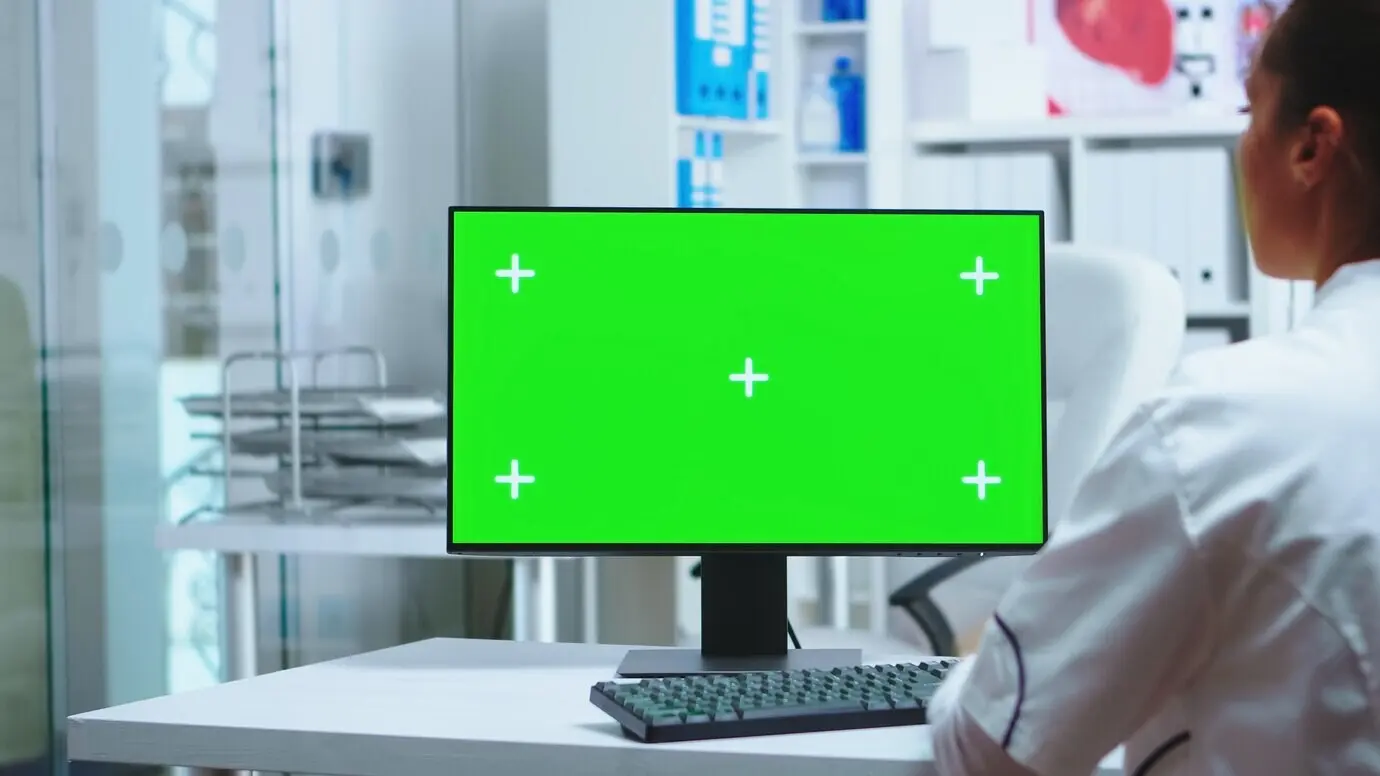 Doctora trabajando en una computadora con pantalla verde en el consultorio de una clínica privada. Asistente con uniforme. Médica con bata blanca usando un monitor con croma en el consultorio para verificar el diagnóstico de un paciente.