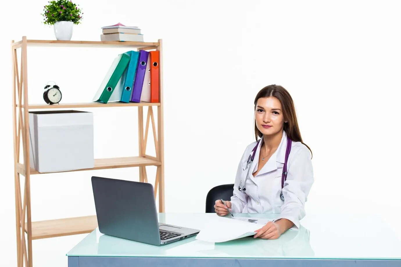 Médico con portátil sentado en un consultorio médico, redactando un informe y sonriendo, aislado frente a una pared blanca.